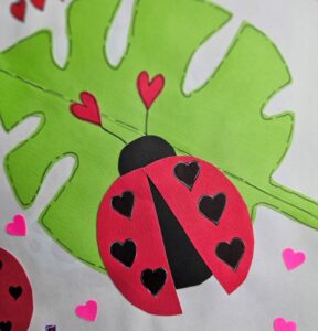 A colorful paper craft of a ladybug with heart-shaped spots on a green leaf, surrounded by small heart shapes.